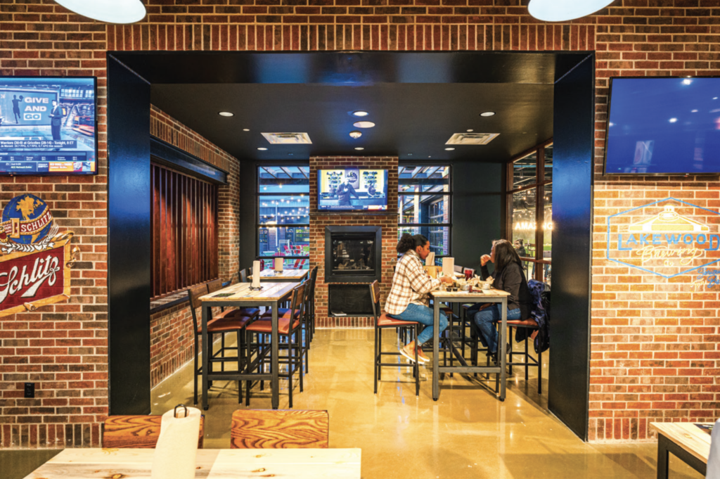 Two people sit at a high table inside a cozy, brick-walled restaurant with sports decor, a TV above a fireplace, and large windows showing more seating and lights in the background.