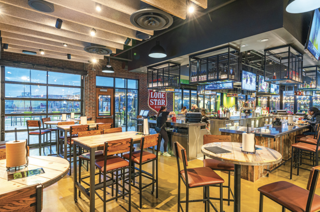 A brightly lit sports bar with wooden tables and chairs, large windows, multiple TV screens, and a bar area. A Lone Star Beer sign hangs near the bar, and a few people are gathered by the counter.