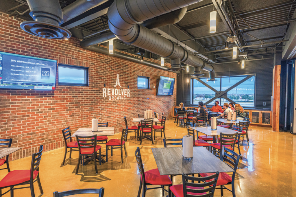Interior of a modern brewery taproom with brick walls, industrial ceiling, and large windows. Empty wooden tables with red and black chairs fill the space; a few people sit near the windows. Several TVs are mounted on the walls.