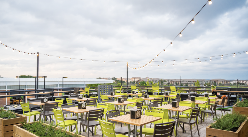 A rooftop patio with many green and brown chairs around wooden tables, string lights overhead, and a cloudy sky in the background. The space overlooks buildings and a distant view of a city or suburb.