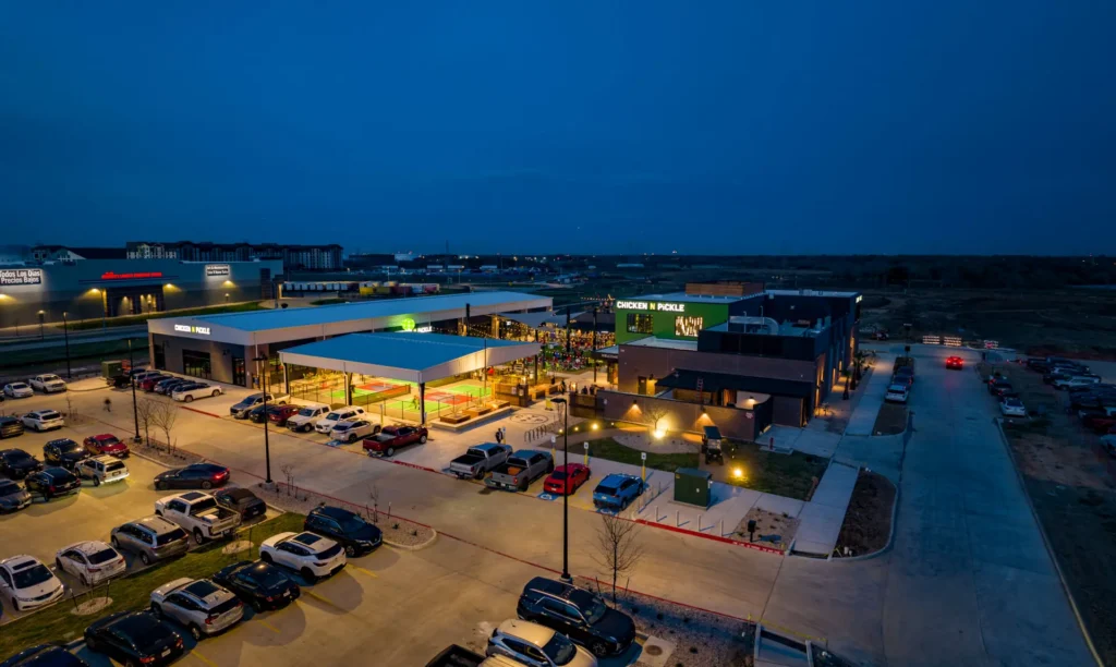 A brightly lit shopping center at dusk in Webster features a parking lot with many cars and several storefronts, including a “Chicken N Pickle” restaurant, surrounded by other buildings and a mostly empty road.