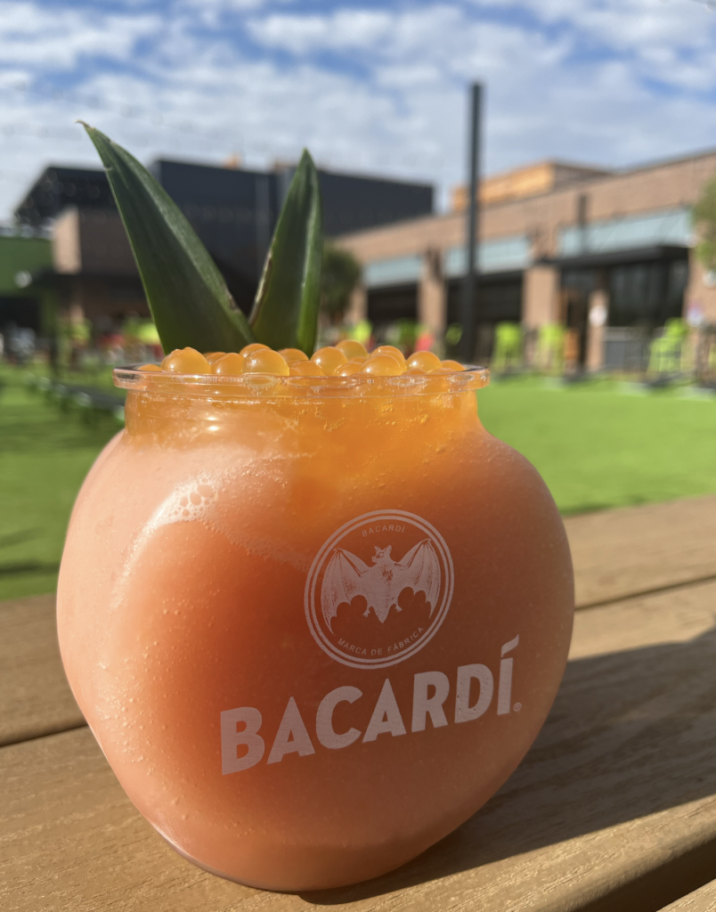 A round Bacardi-branded glass filled with a pink frozen cocktail, topped with orange pearls and pineapple leaves, sits on a wooden table outdoors with a blurred green lawn and modern buildings in the background.