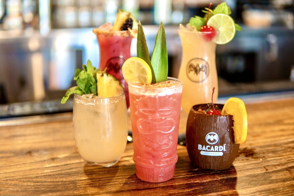 Four colorful tropical cocktails garnished with fruits, mint, and umbrellas are displayed on a wooden bar. The drinks include a Bacardi cup, tiki glass, pale yellow drink with pineapple, and a pink drink with lemon and pineapple leaves.