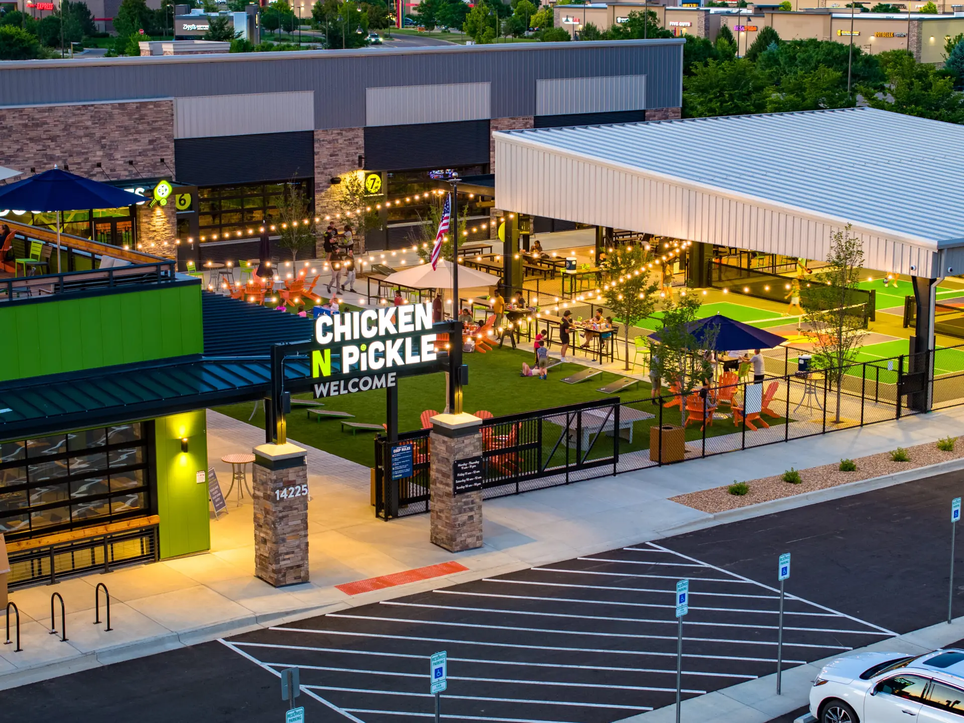 A lively outdoor venue in Thornton with a Chicken N Pickle Welcome sign, string lights, pickleball courts, seating areas, and people playing games, surrounded by modern buildings and a parking lot with accessible spaces.