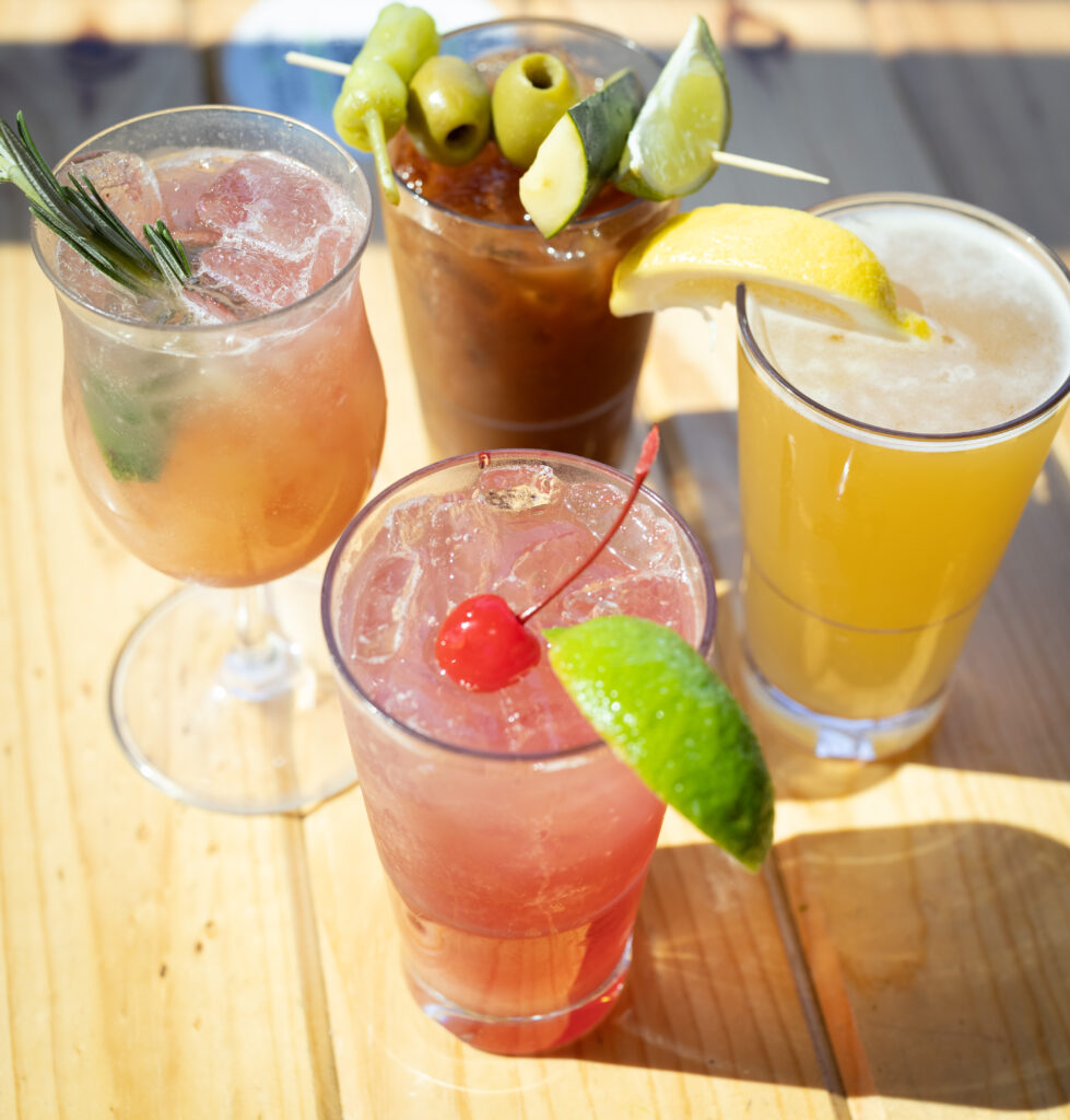 Four colorful cocktails on a wooden table, each garnished with fruit or herbs: a pink drink with a cherry and lime, a yellow drink with a lemon wedge, an orange drink with rosemary, and a dark drink with olives and lime.