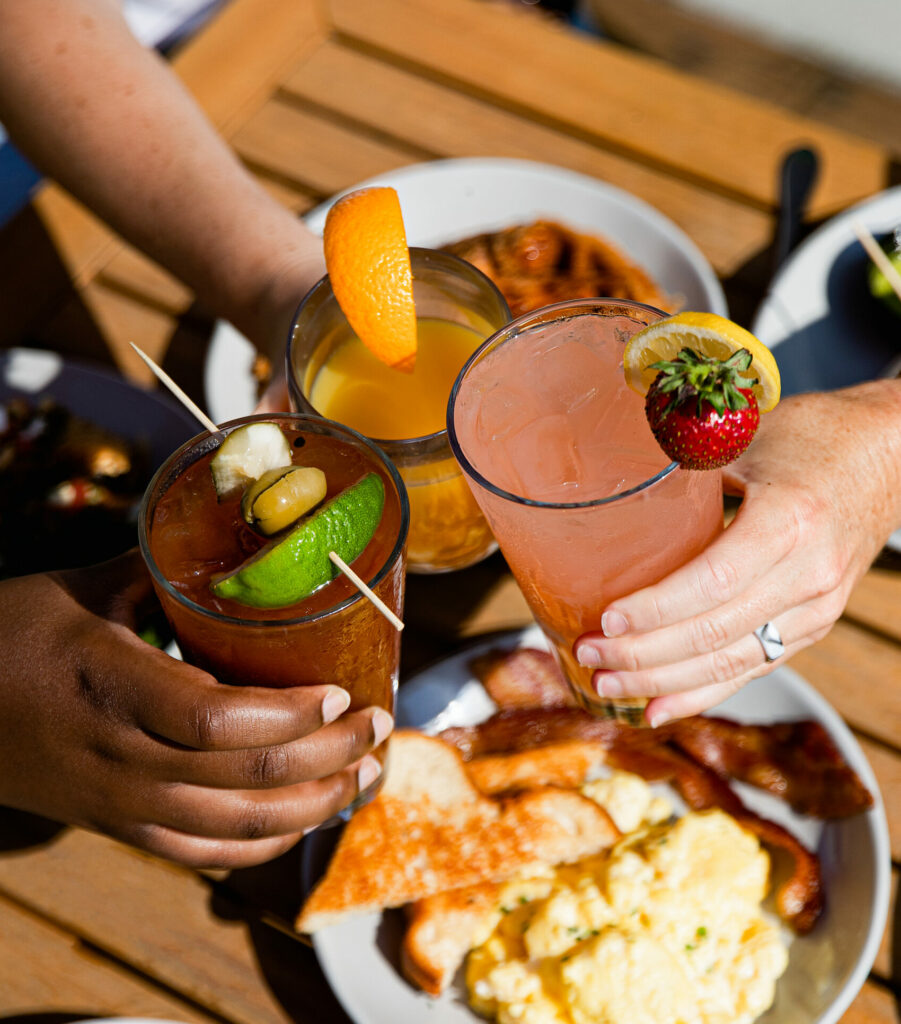 Three hands toast with colorful cocktails over a wooden table set with breakfast plates, including scrambled eggs, bacon, toast, and fruit. One drink is garnished with a lime, another with orange, and another with a strawberry and lemon.