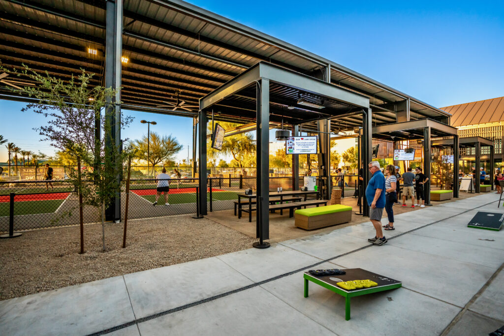 People play pickleball on outdoor courts with shaded seating areas nearby. Others are walking and playing cornhole on a sunny day. The scene is lively, with clear blue skies and desert plants around.
