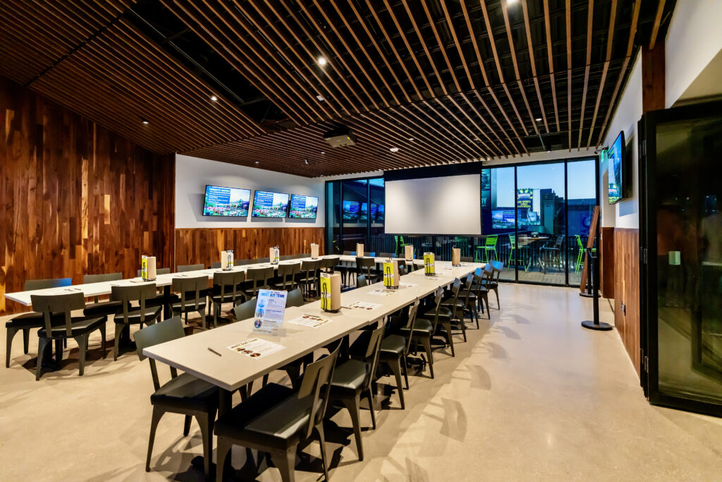 A modern, well-lit meeting room with long tables and black chairs, large screen displays on the wall, condiments on each table, wood accents, and glass doors opening to an outdoor patio.
