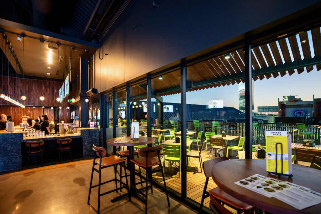 A modern bar interior with high tables and chairs, a view of an outdoor patio with green chairs and tables through large glass windows, and people gathered near the bar counter in the background.