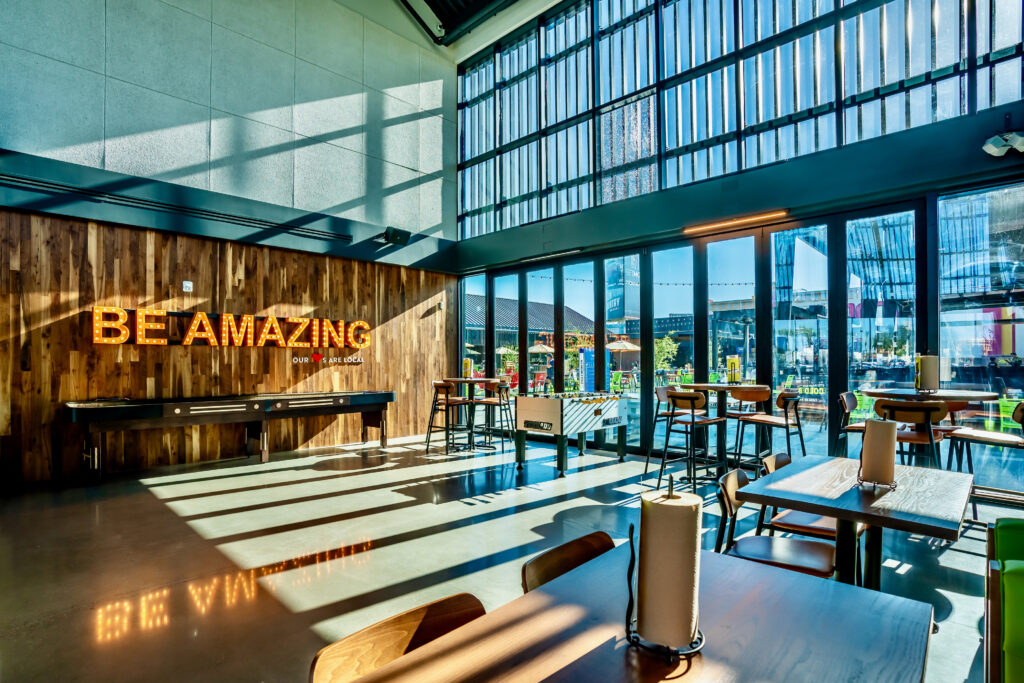 A modern, sunlit cafe with large windows, wooden walls, and a neon sign that says BE AMAZING. Tables and high stools are arranged throughout, with outside seating visible through the glass.