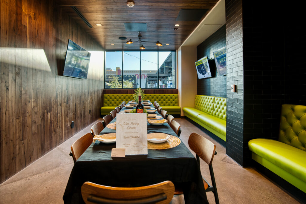 A modern dining room with a long table set for a meal, surrounded by wooden chairs and bright green cushioned benches along the walls. Large windows let in natural light, and two TVs are mounted on the walls.