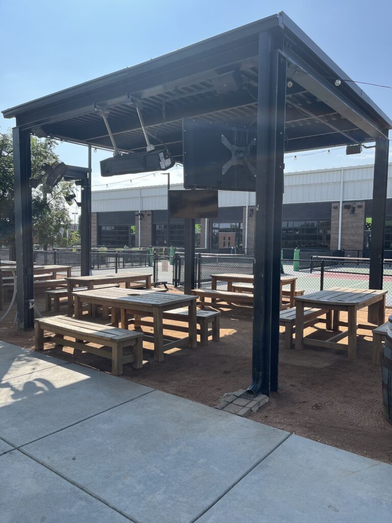 A shaded outdoor seating area with wooden picnic tables and benches under a black metal pergola, located next to a fenced tennis or pickleball court on a sunny day.
