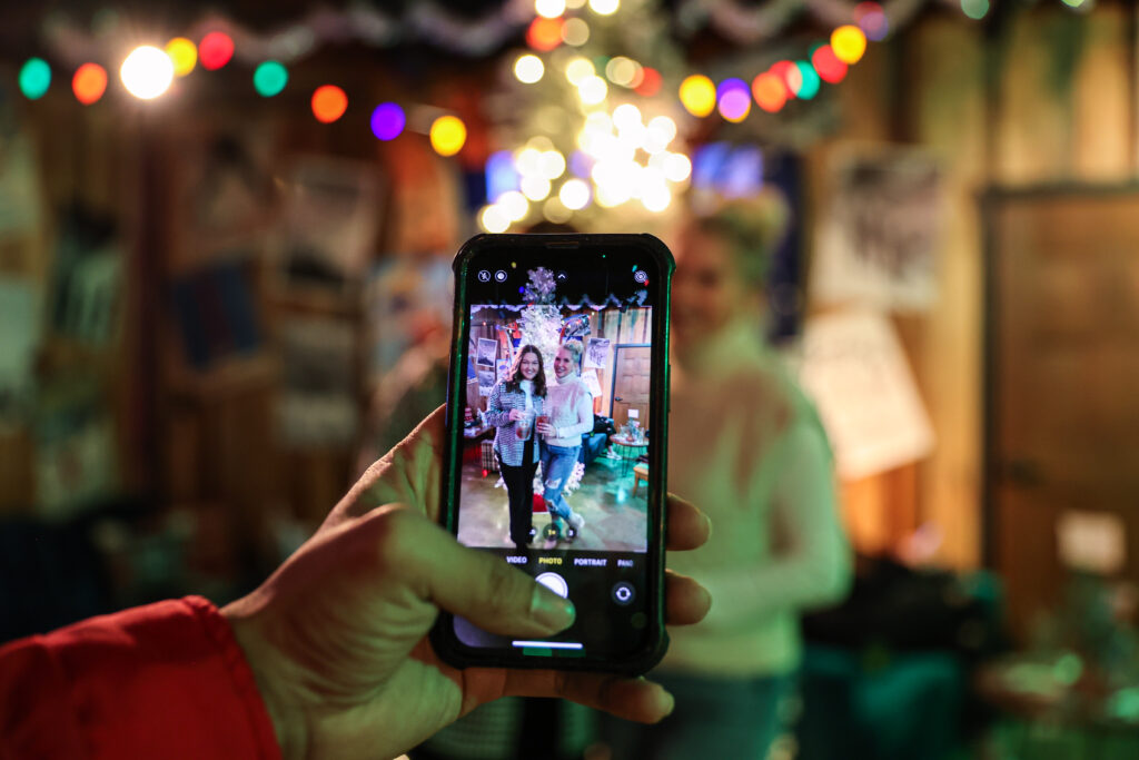 A hand holds a phone, capturing a photo of two people posing together in a festive, colorful room with string lights and decorations in the background. The phone screen shows the clear image, while the background is blurred.
