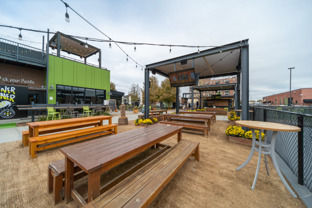 An outdoor patio area with long wooden picnic tables, a few high-top tables, string lights overhead, and yellow potted flowers, surrounded by modern buildings and a cloudy sky.