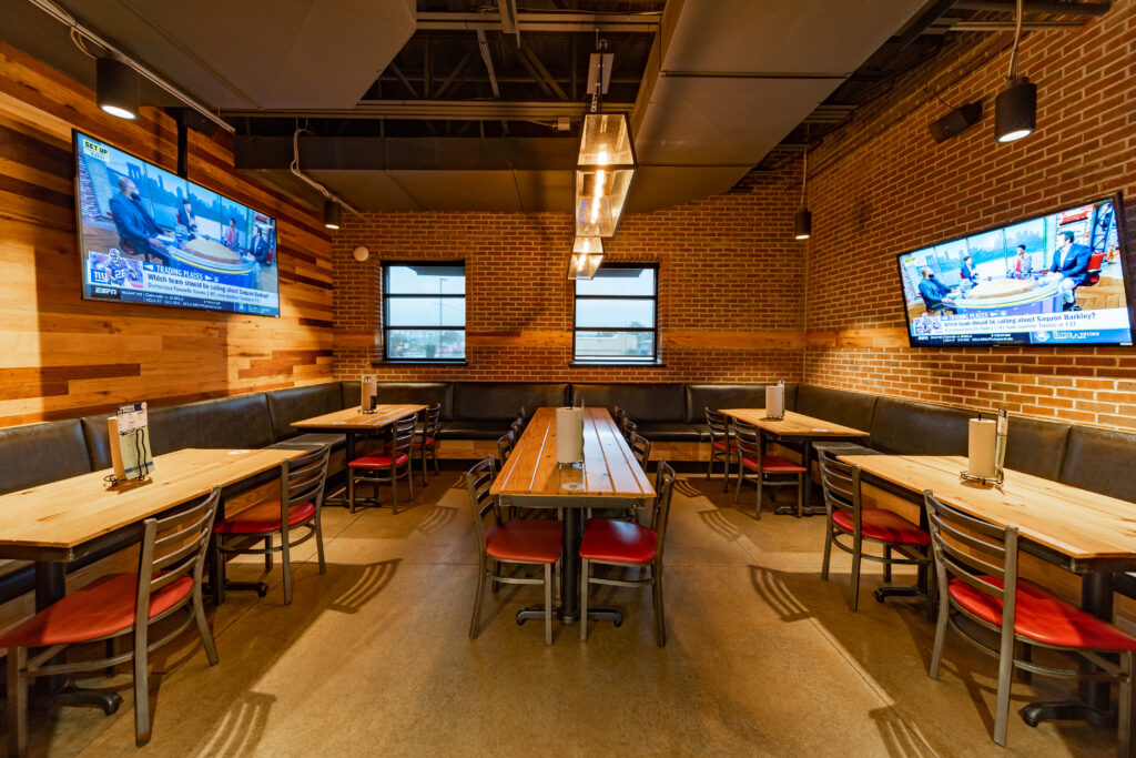 A modern restaurant dining area with wooden tables, red chairs, brick and wood-paneled walls, two wall-mounted TVs, black cushioned benches, and large windows letting in natural light.