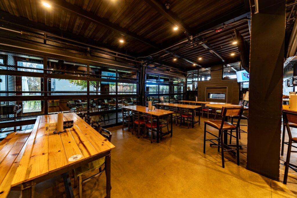 Spacious modern restaurant with wooden tables and metal chairs, polished concrete floor, large windows, and exposed ceiling beams. The area appears empty and well-lit with natural and overhead lighting.