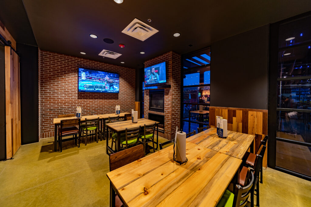 A modern restaurant dining area with brick walls, wooden tables, and chairs. Two flat-screen TVs are mounted on the wall, showing sports. The space is well-lit with a relaxed atmosphere.