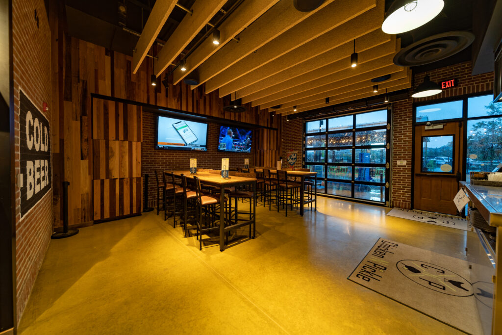 A modern, empty restaurant interior with wooden tables and high chairs, large wall-mounted TVs, wood and brick walls, large windows, and a Cold Beer sign on the left. Warm lighting creates a cozy atmosphere.