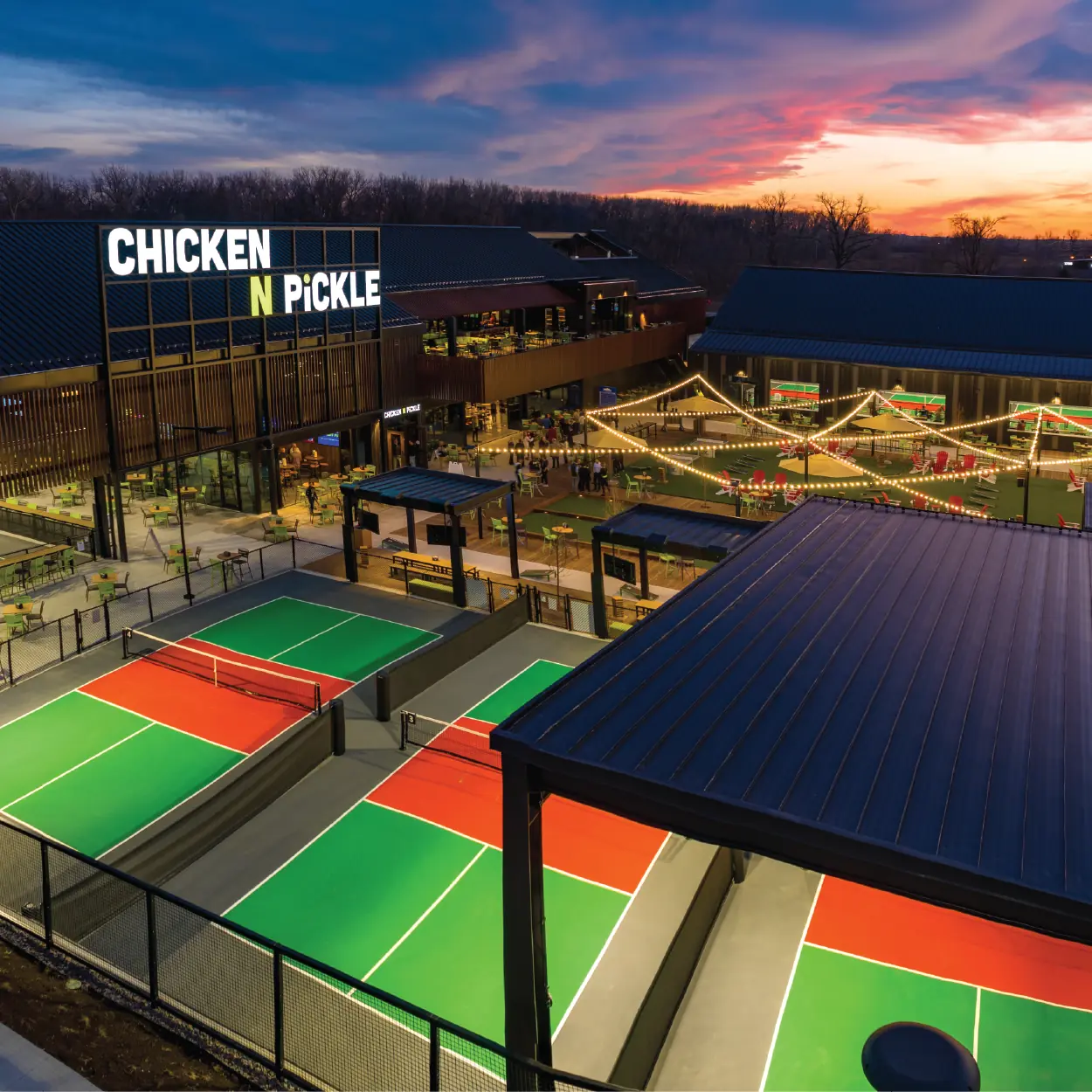 Aerial view of Chicken N Pickle at sunset, showing brightly lit pickleball courts, outdoor seating, string lights, and a modern building with a sign reading “CHICKEN N PICKLE.”.