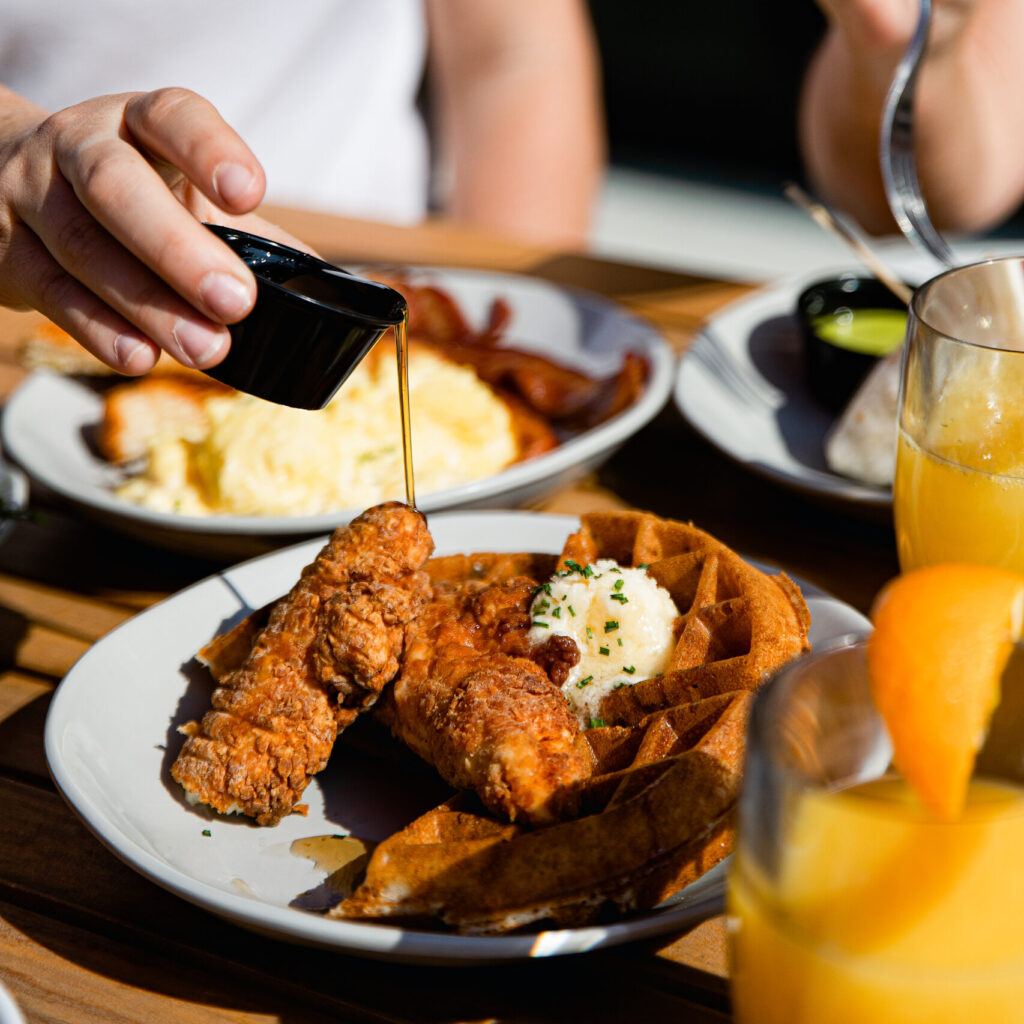 A person pours syrup over fried chicken and waffles on a plate, served with butter and garnish—perfect for an Easter weekend brunch. Scrambled eggs, bacon, and a glass of orange juice are visible on the table.