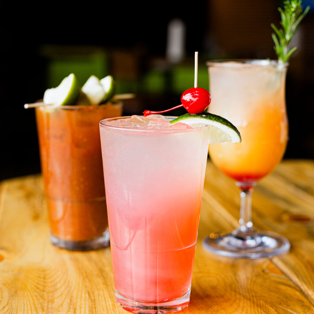 Three colorful cocktails on a wooden table: a pink drink garnished with a cherry and lime, a reddish drink with lime wedges, and an orange-yellow drink with a rosemary sprig in a curvy glass.