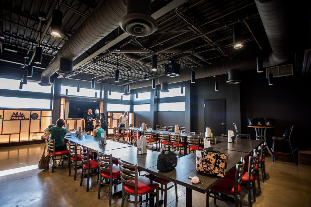 Modern meeting room with long tables, red-cushioned chairs, exposed ceiling pipes, and several people gathered near a screen at the far end. Large windows let in natural light.