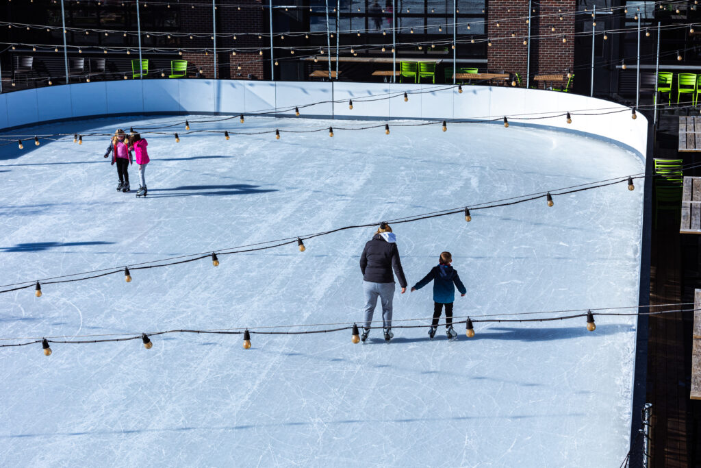 People ice skating on an outdoor rink under string lights, with a few groups scattered across the ice. Green chairs are visible outside the rink near a brick building in the background.