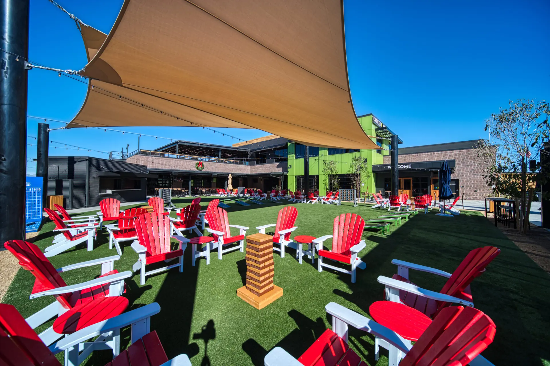 Outdoor seating area in Henderson featuring numerous red Adirondack chairs on artificial grass, a large Jenga game at the center, sun shades overhead, and modern buildings in the background beneath a clear blue sky.
