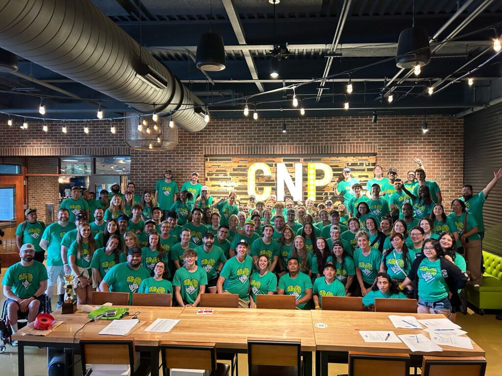 A large group of people wearing matching green shirts pose and smile together inside a well-lit room with brick walls and a sign reading CNP in large letters. Tables with papers are in the foreground.