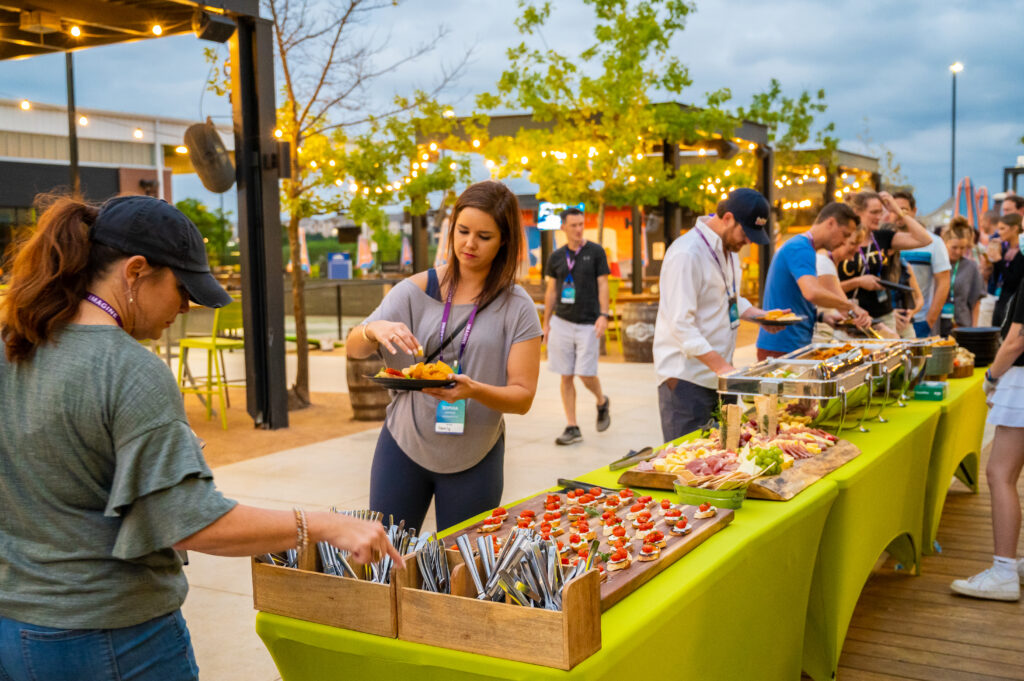People serve themselves food from a long buffet table outdoors at a casual event. The table is covered with a green tablecloth and a variety of dishes. String lights and trees decorate the lively setting.