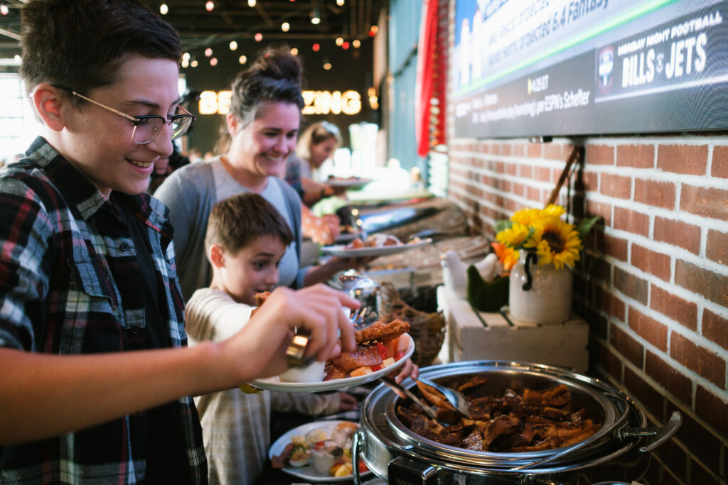 A group of smiling people, including children, enjoy an Easter weekend brunch, serving themselves food from a buffet table in a warmly lit restaurant with brick walls and string lights—perhaps after a fun egg hunt.