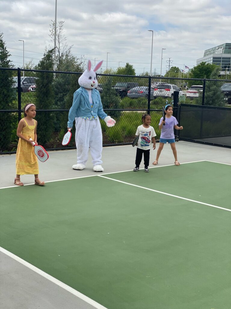 Three children stand on a tennis court holding racquets next to a person in an Easter Bunny costume, enjoying tennis outdoors on a cloudy day—perhaps after an Easter weekend egg hunt.