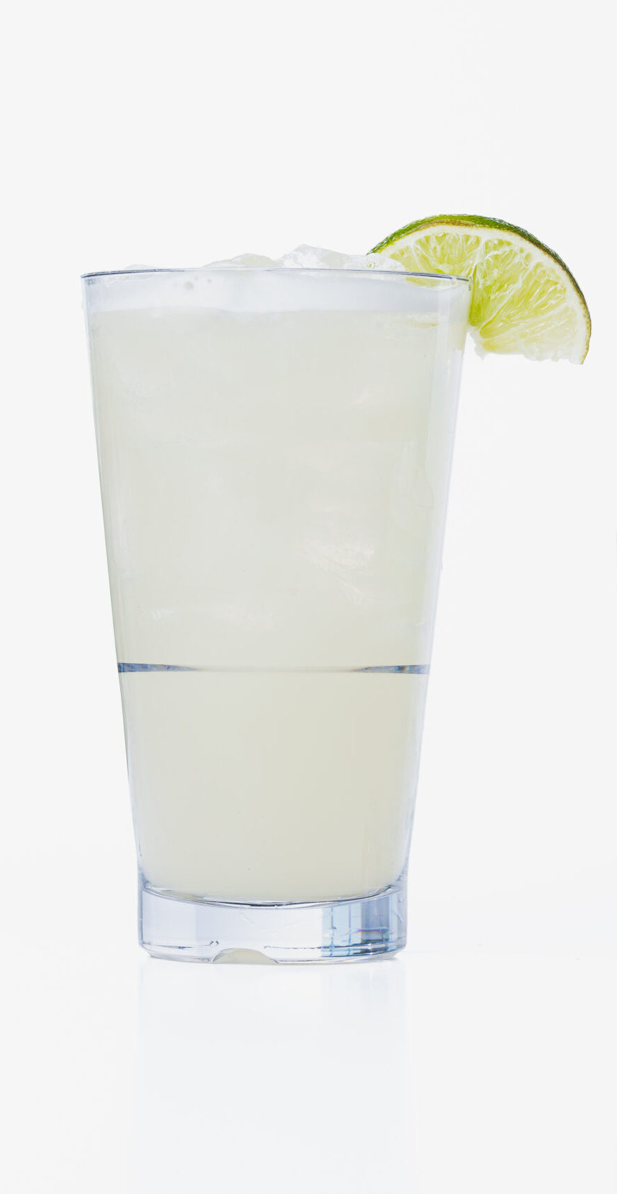 Two glasses filled with a light yellow drink, each topped with ice and garnished with a lime wedge, sit side by side against a white background.