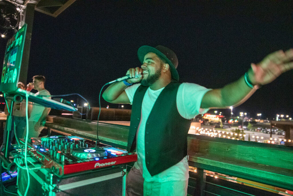 A DJ wearing a hat, white shirt, and black vest performs energetically with a microphone at an outdoor nighttime Easter weekend brunch, with DJ equipment and city lights visible in the background.