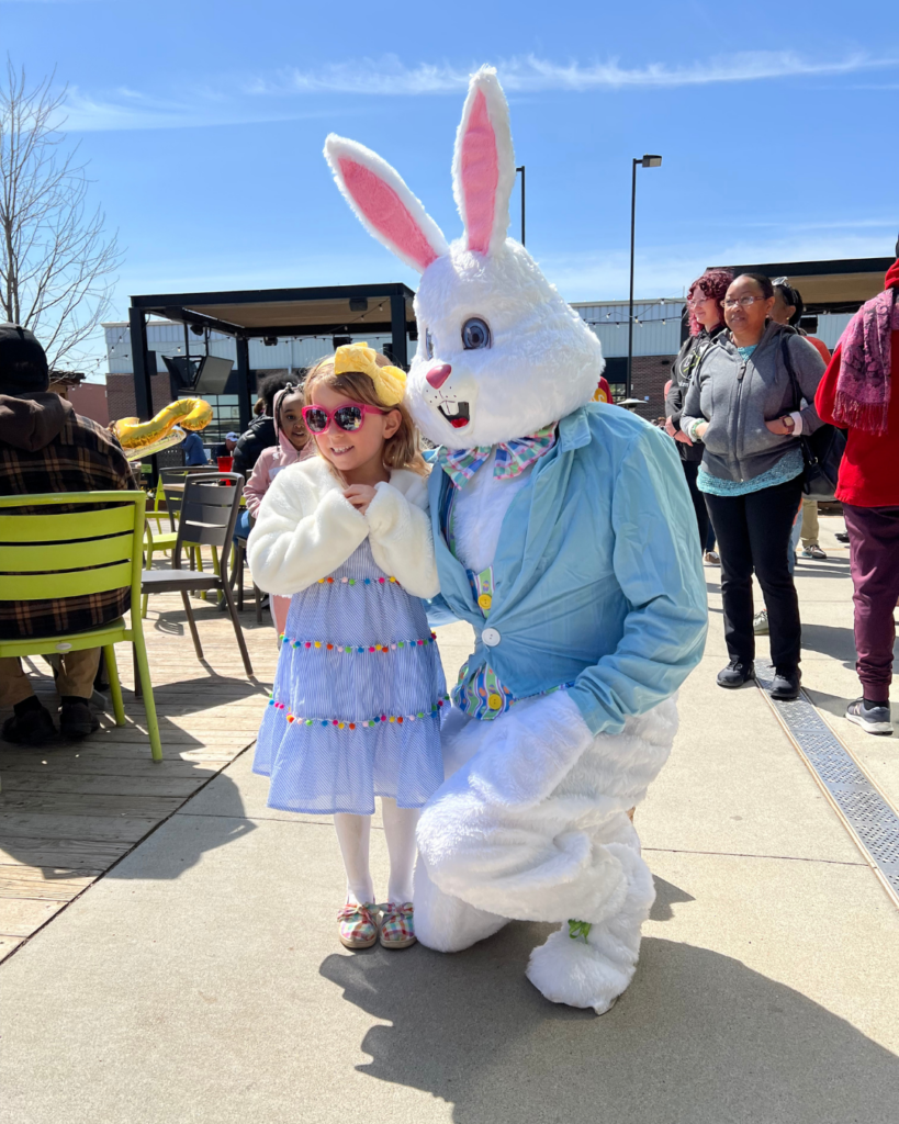 A young girl in a blue dress and heart-shaped sunglasses poses with a person in a large Easter Bunny costume at an outdoor event on a sunny day, enjoying the Easter weekend egg hunt surrounded by other people.