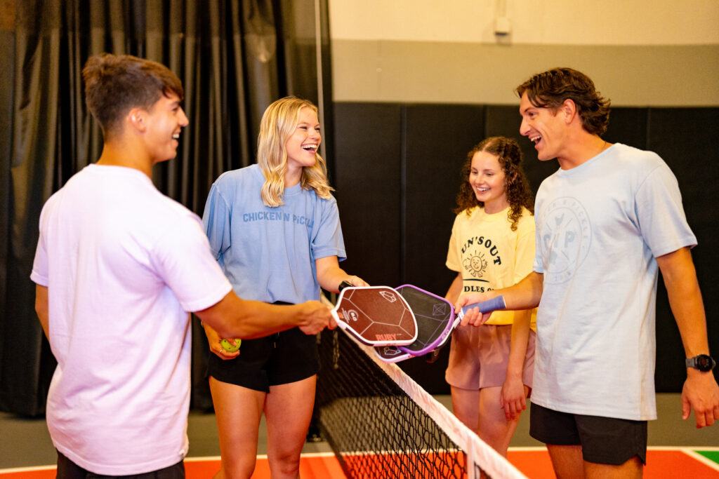 Four young adults stand on an indoor pickleball court, smiling and touching paddles over the net, appearing cheerful and ready to play. Two hold pickleball paddles and one holds a ball.
