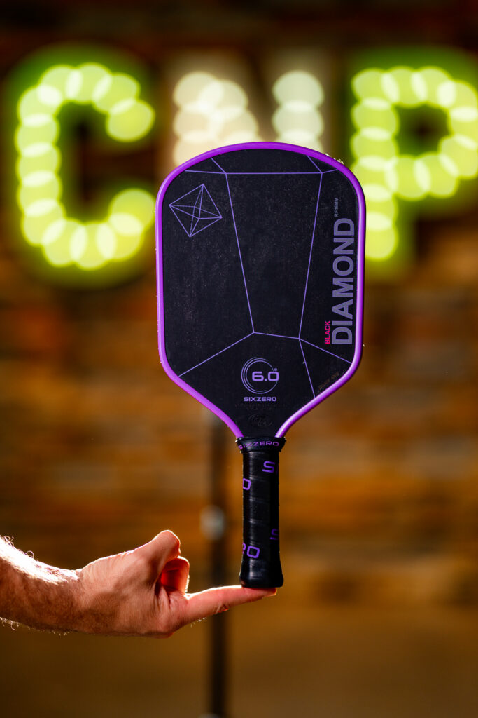 A person balances a black and purple pickleball paddle labeled “Black Diamond” on their index finger, with blurred yellow-green lights and a brick wall in the background.