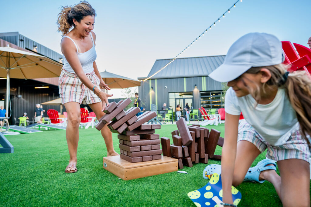 A woman and a young girl play an outdoor giant Jenga game on green grass during an Easter weekend brunch. The woman laughs as the blocks fall, while the girl crouches nearby. People, umbrellas, and modern buildings are visible in the background.