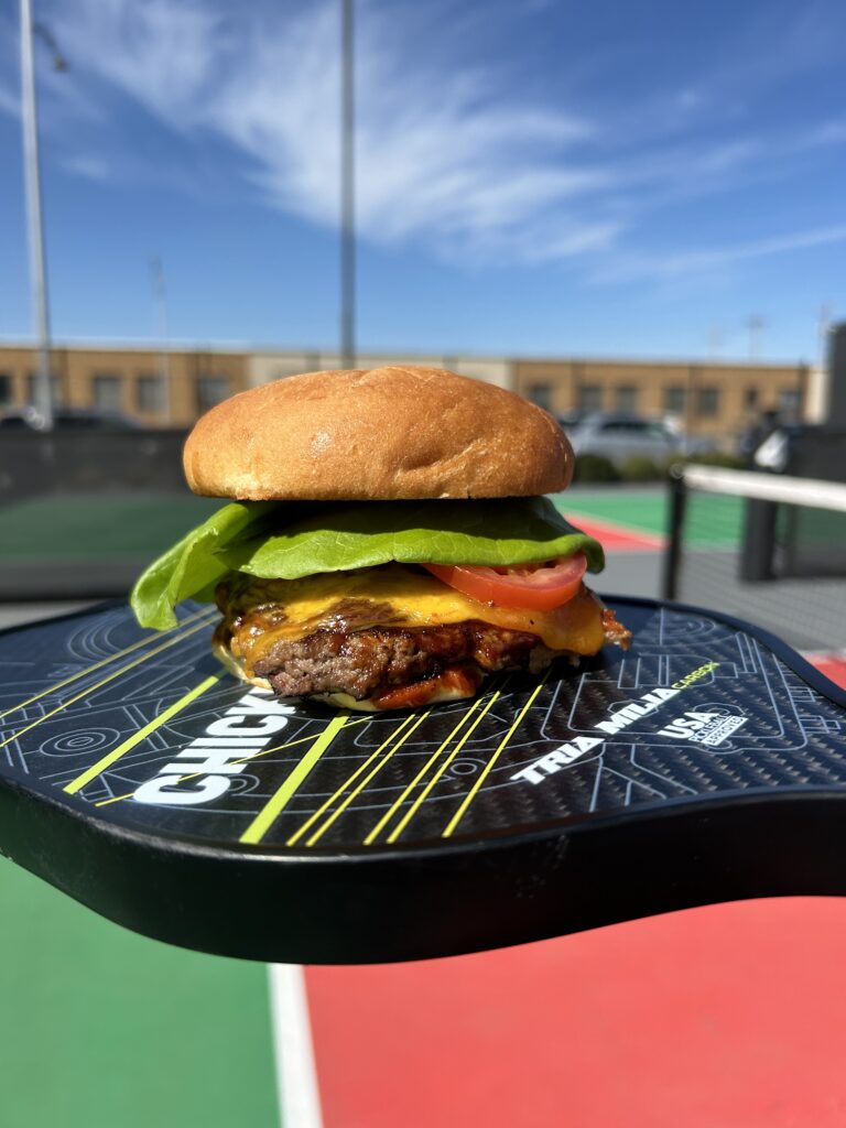 A cheeseburger with lettuce and tomato sits on a pickleball paddle, outdoors on a court, ready for an Easter weekend brunch or egg hunt, with a bright blue sky and buildings in the background.