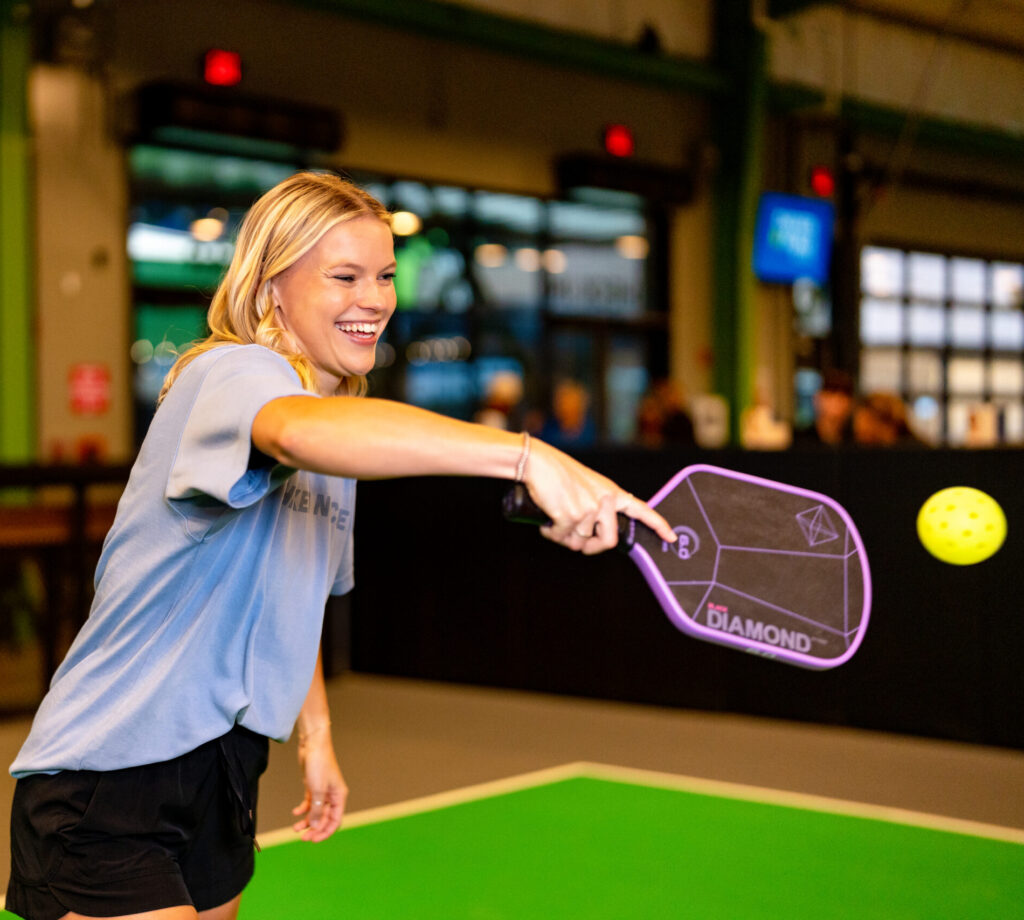 A woman wearing a light blue shirt and black shorts smiles as she plays pickleball indoors, reaching out to hit a yellow ball with a purple paddle.
