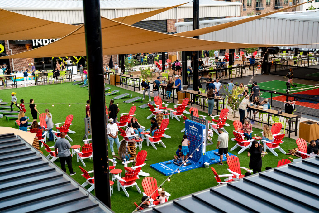 A lively outdoor space with people socializing, playing games, and sitting in red Adirondack chairs on artificial grass, surrounded by tables and shaded by tan canopies, next to modern buildings.