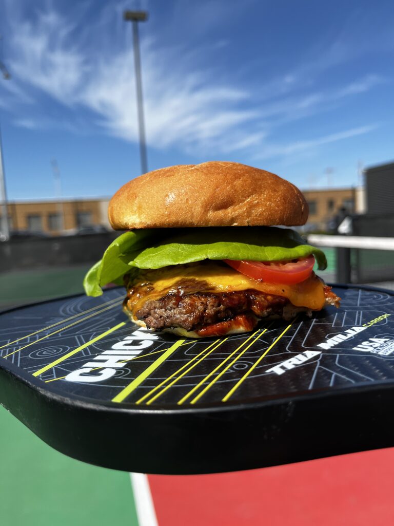 A cheeseburger with lettuce, tomato, and cheddar cheese sits on a pickleball paddle against an outdoor court backdrop under a bright blue sky, perfect for an Easter weekend brunch after the egg hunt.