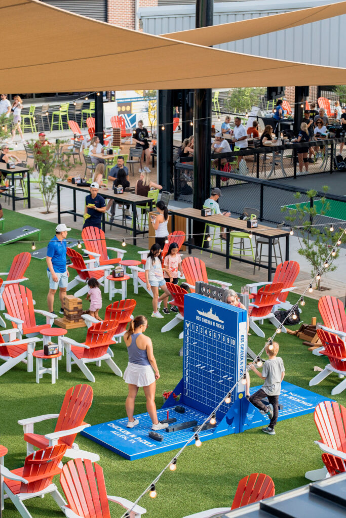 People enjoy an outdoor space with red and white Adirondack chairs, playing oversized games like Connect Four and Jenga on artificial grass, while others sit at tables under canopies. String lights hang above the area.