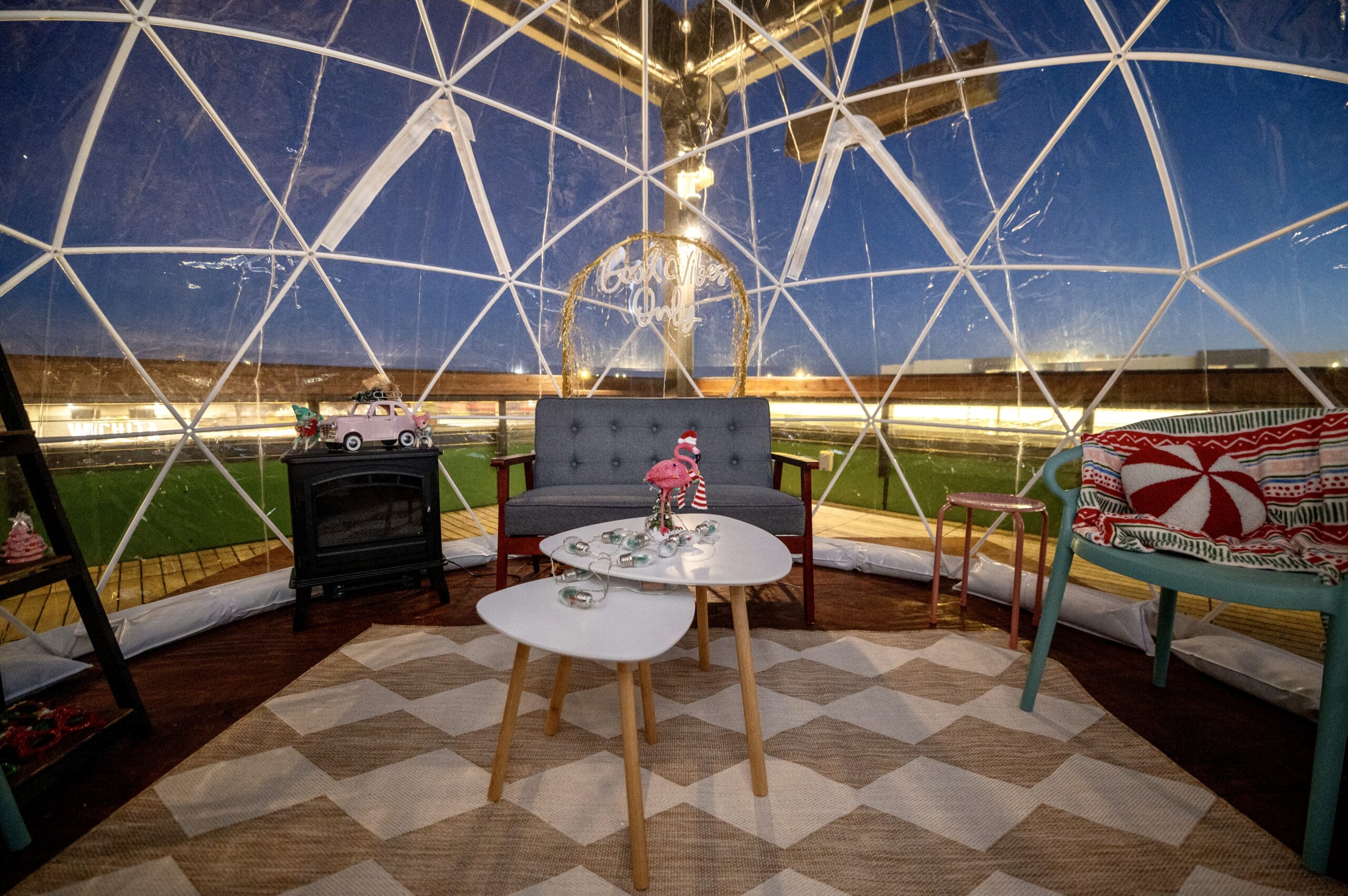 A cozy seating area inside a transparent dome features a gray sofa, a blue bench with festive pillows, a fireplace, and two white coffee tables on a checkered rug, all surrounded by evening outdoor scenery.