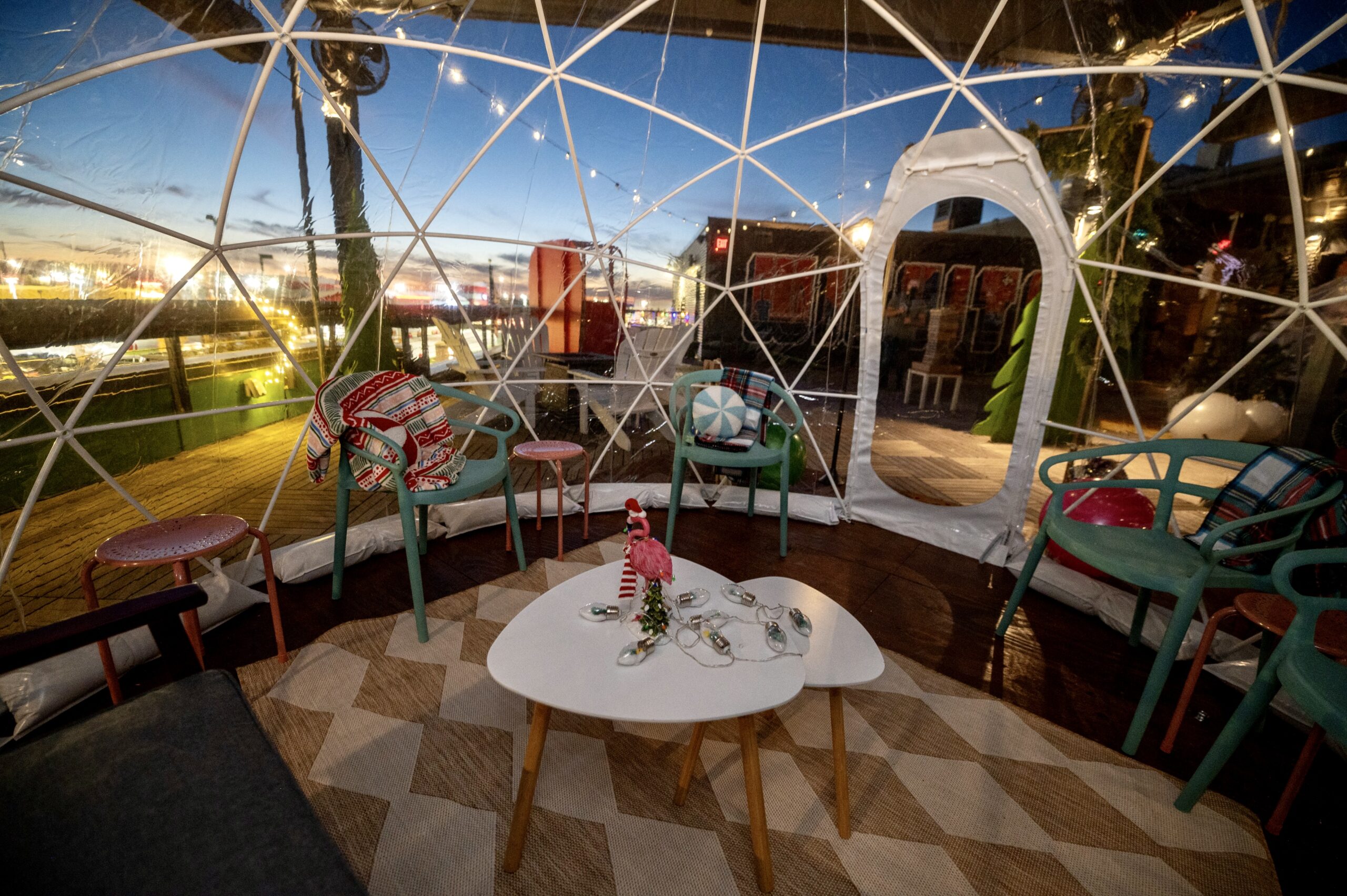 A cozy seating area inside a transparent dome with colorful chairs, a patterned rug, and a white table with cups. String lights hang above, and city lights are visible outside at dusk.