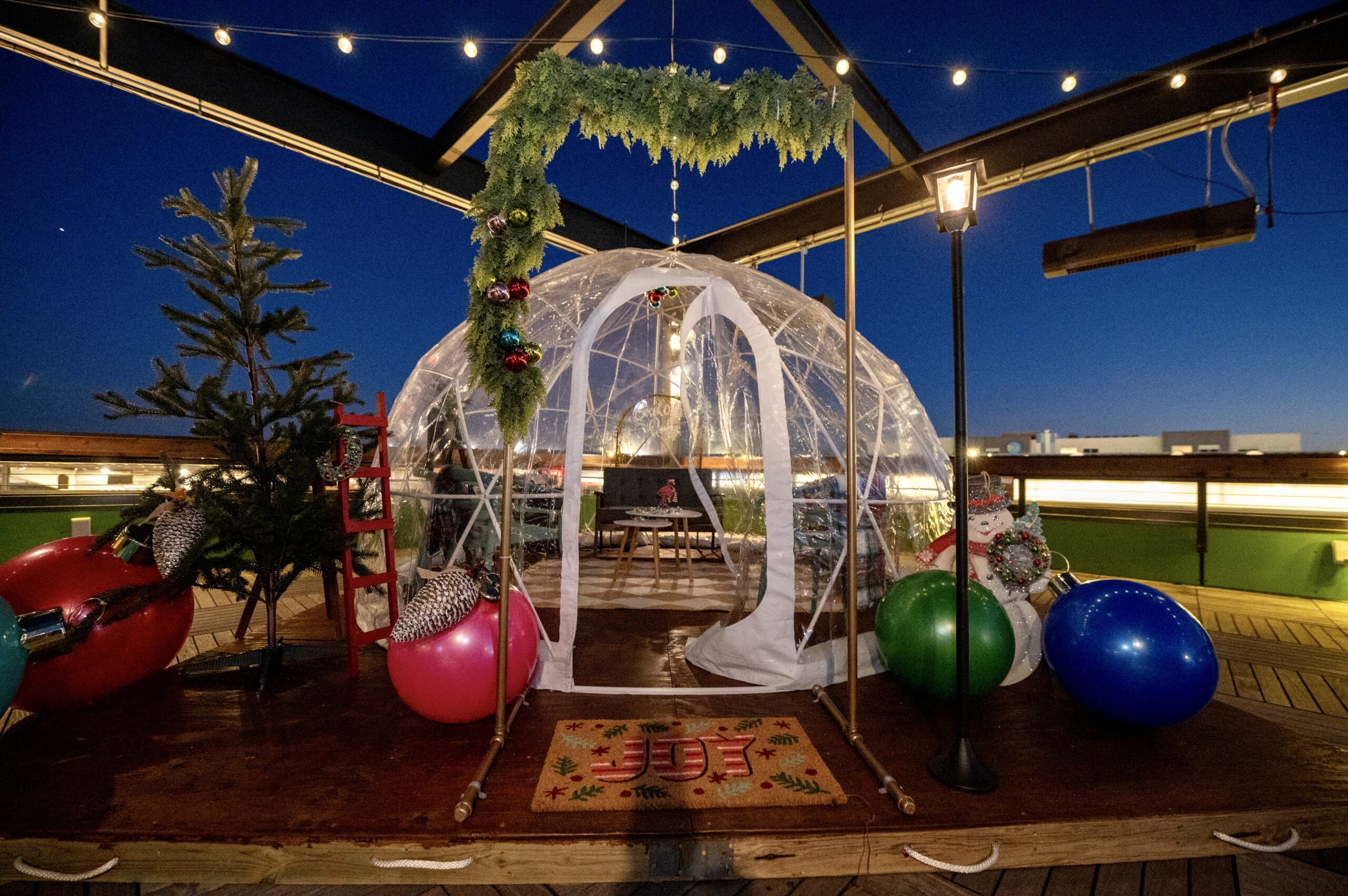 A transparent dome tent decorated with festive greenery stands on a rooftop deck with string lights, large colorful ornaments, a small Christmas tree, and a holiday doormat at dusk.