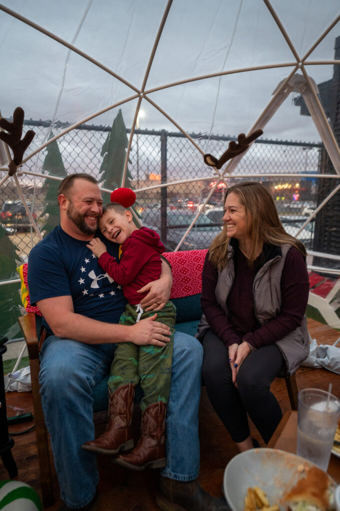 A man, woman, and young boy sit together inside a clear dome, smiling and laughing. The boy wears reindeer antlers and cowboy boots while sitting on the mans lap. The setting appears festive and cozy.