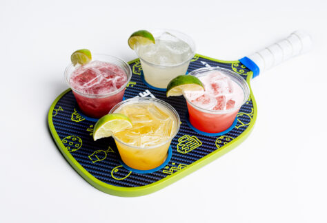 Pickleball Paddle holding four small margraitas with limes, Mango, Classic, Spicy Blackberry, & Strawberry flavors