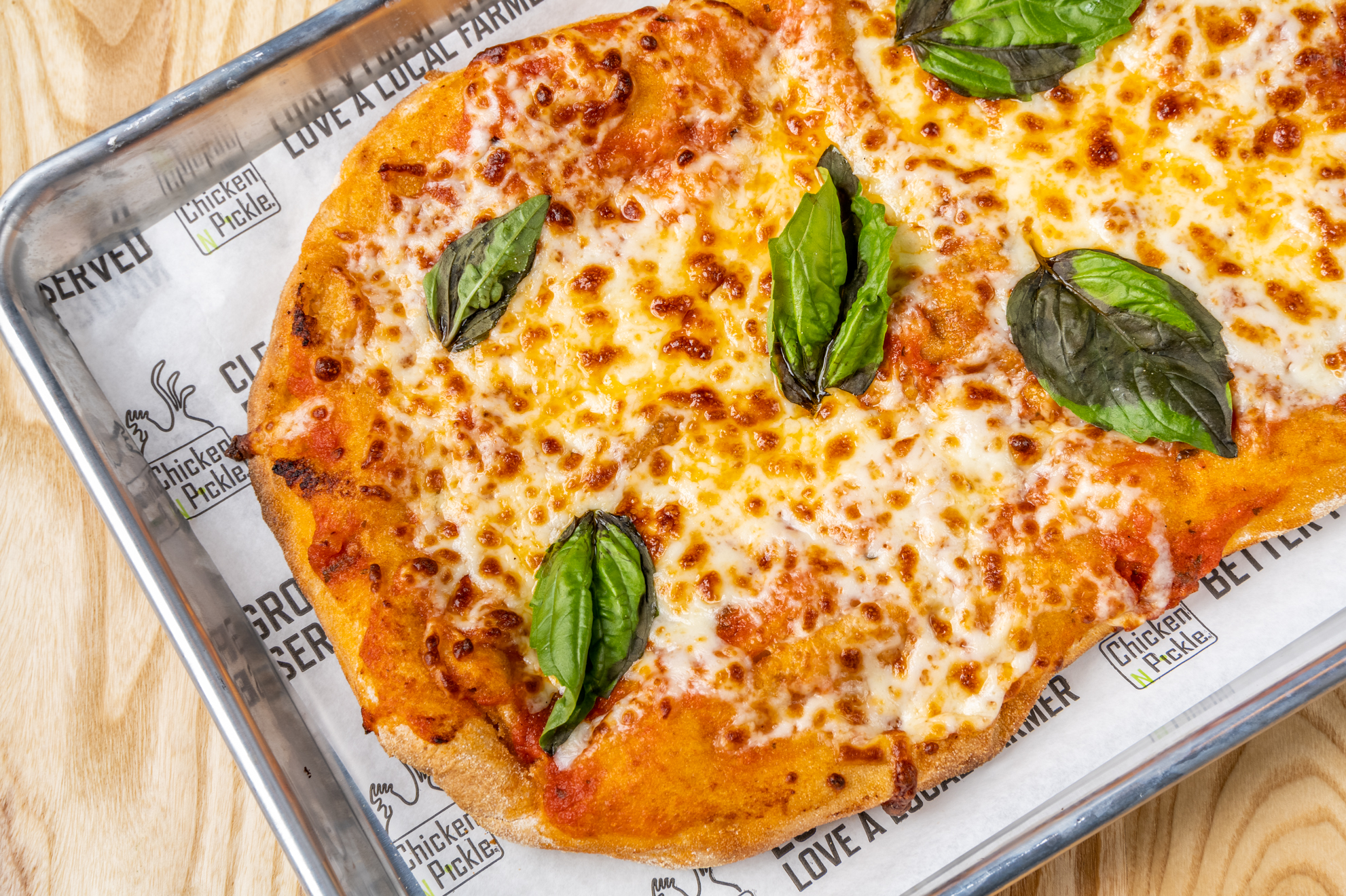 A rectangular margherita pizza topped with melted cheese, tomato sauce, and fresh basil leaves, served on a metal tray lined with branded paper.