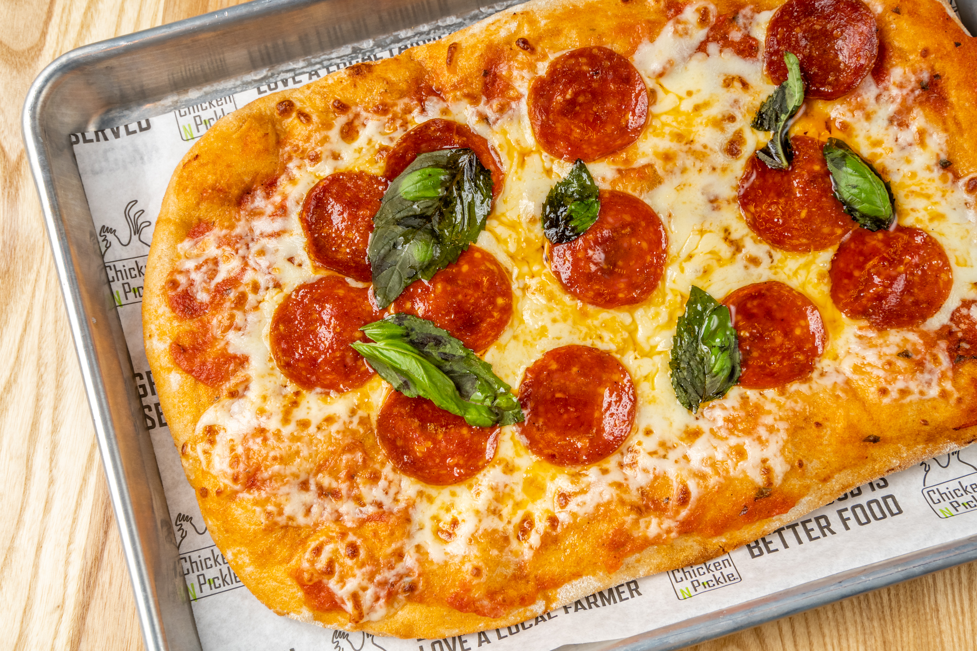 Rectangular pepperoni pizza on a metal tray, topped with melted cheese, pepperoni slices, and fresh basil leaves, resting on branded parchment paper, on a wooden table.